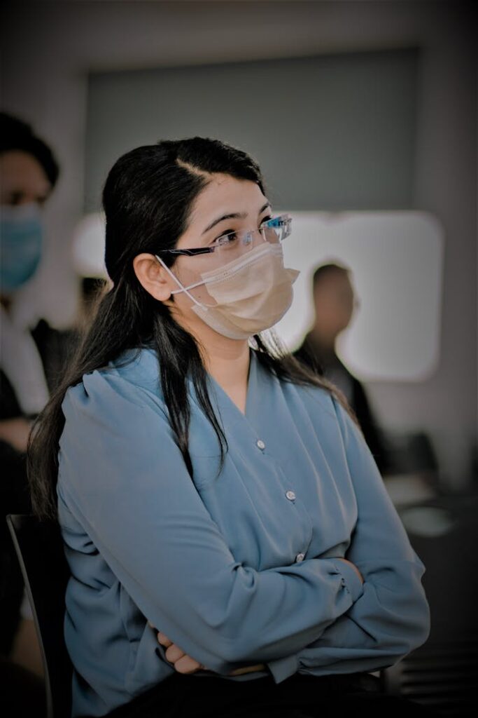 A woman in a blue blouse wearing eyeglasses and a face mask, attentively sitting indoors.