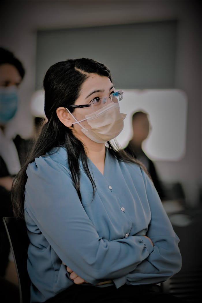 A woman in a blue blouse wearing eyeglasses and a face mask, attentively sitting indoors.