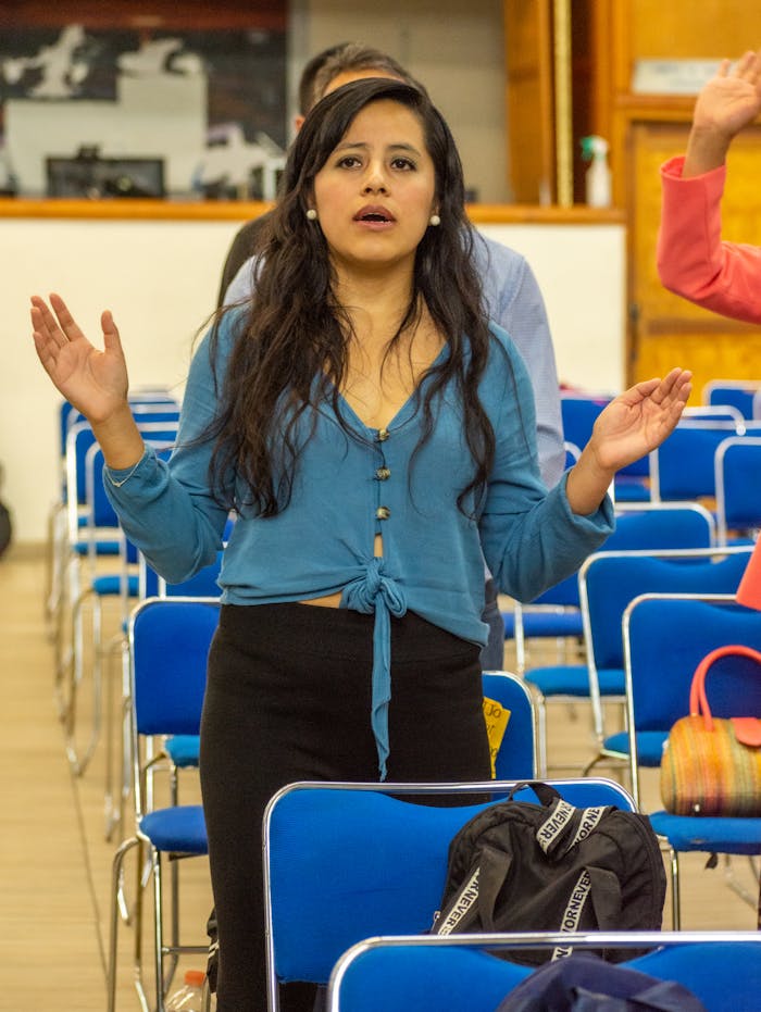 A woman with long hair stands with raised hands during an indoor religious event.
